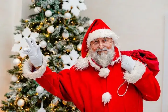 Moș Crăciun salutând cu mâna, cu sacul pe umăr, lângă bradul cu globuri albe și luminițe